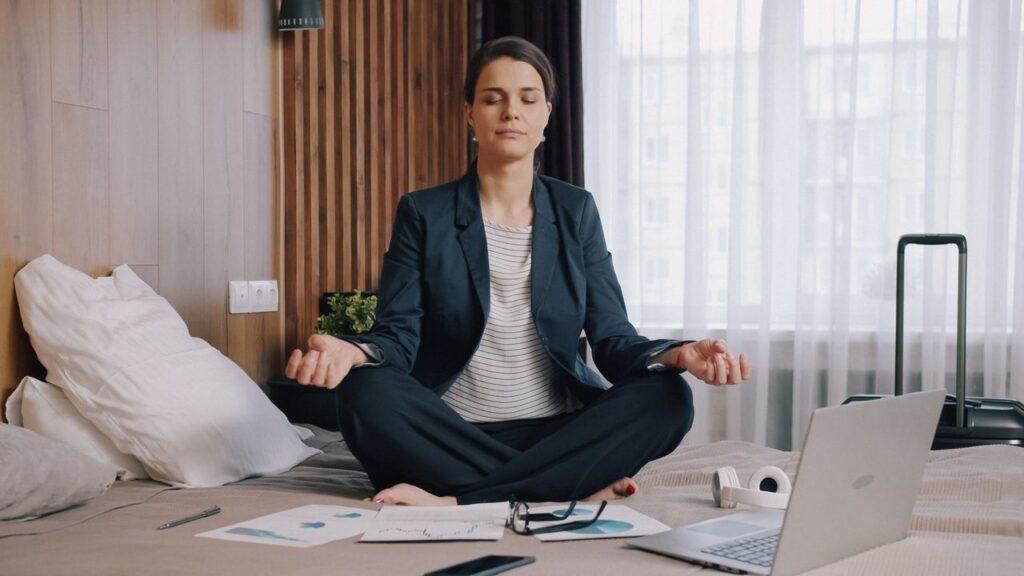 Betriebliche Gesundheitsförderung: Warum Zeitmangel ein Designproblem ist Frau im Business-Outfit meditiert im Hotelzimmer zwischen Koffer und Laptop